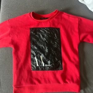 Röd sweatshirt från Armani Exchange - Cool röd sweatshirt från Armani Exchange med stort svart tryck framtill där det står 'A|X BEATS'. Tröjan har rund halsringning och långa ärmar. Perfekt för dig som gillar streetstyle och vill sticka ut med en snygg logga.