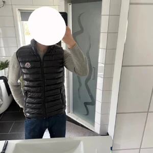 Svart dunväst från Moncler - Snygg svart dunväst från Moncler med glansig finish och horisontella quiltade sömmar. Västen har hög krage, dragkedja framtill och Moncler-logga på bröstet. Perfekt att slänga över en hoodie eller tröja för en clean look.