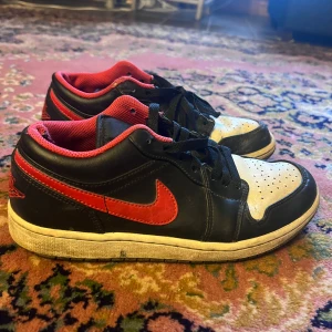 Nike Air Jordan 1 Low svart/röd/vit - Säljer ett par Nike Air Jordan 1 Low sneakers i svart, rött och vitt skinn. Klassisk siluett med röd swoosh, svart ovandel och vit tåbox. Röd insida och broderad Air Jordan-logga på hälen. Perfekta för dig som gillar streetwear och ikoniska sneakers.