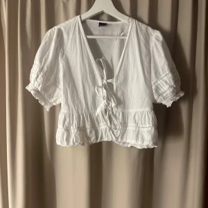Vit blus med knyt och volang - Söt vit blus med korta puffärmar, v-ringning och knyt framtill. Blusen har volangdetaljer längs nederkanten och ärmarna samt fina broderade inslag.💓