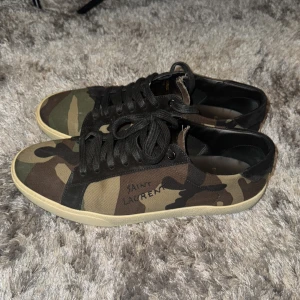 Saint Laurent camo sneakers - Säljer ett par Saint Laurent sneakers i klassisk camo med svarta detaljer och snörning. Skorna har en låg profil, rund tå och vit sula. Tillverkade i canvas med läderdetaljer och Saint Laurent-logga på sidan. Perfekta för dig som vill sticka ut med en cool streetstyle.