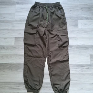 Olivgröna cargopants med snörning - Olivgröna cargopants med elastisk midja och grön snörning. Byxorna har stora sidofickor med lock, resår vid benslut och är tillverkade i ett slitstarkt material. Perfekta för en avslappnad streetstil.