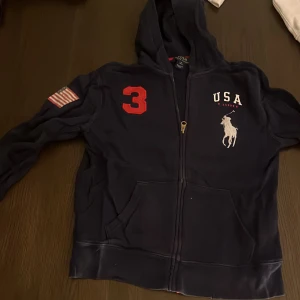 Marinblå hoodie Polo Ralph Lauren - Marinblå hoodie från Polo Ralph Lauren med dragkedja, stor vit logga och USA-tryck på bröstet. Röd siffra 3 på axeln och amerikansk flagga på ärmen. Klassisk huva och kängurufickor. Perfekt för dig som gillar sportig och stilren look.