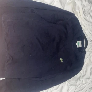 Mörkblå sweatshirt från Lacoste - Klassisk mörkblå sweatshirt från Lacoste med rund halsringning och den ikoniska gröna krokodilloggan broderad på bröstet. Tröjan är unisex och har långa ärmar samt ribbade muddar vid ärmslut och nederkant. Perfekt för en clean och stilren look.