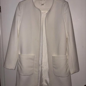 Vit strukturerad kappa med fickor - Snygg vit kappa med strukturerat tyg och raka linjer. Jackan har två stora fickor framtill och är öppen utan knäppning. Perfekt för dig som gillar stilrena och minimalistiska plagg. Lätt fodrad och med rundad halsringning.