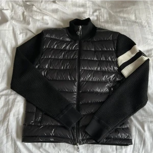 Svart Moncler pufferjacka med stickade ärmar - Snygg svart pufferjacka från Moncler med stickade ärmar och två vita ränder på ena ärmen. Jackan har quiltad front i glansigt material, dragkedja och hög krage. Moncler-logga på ärmen ger en exklusiv vibe. Perfekt för dig som vill ha något unikt och stilrent.
