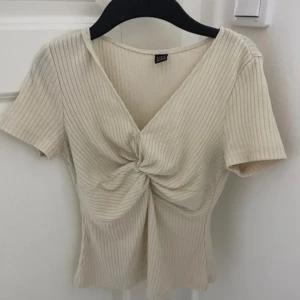 Ribbad beige omlottopp med knut - Superfin ribbad topp i beige från Gina Tricot med kort ärm, v-ringning och snygg omlottdesign med knut framtill. Perfekt till jeans eller kjol för en trendig look. Mjuk och stretchig bomullskänsla.