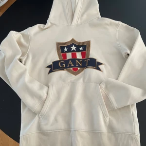 Krämvit hoodie från GANT med emblem - Snygg krämvit hoodie från GANT med stort broderat emblem på bröstet i blått, rött och vitt. Klassisk känguruficka framtill och huva. Perfekt för dig vill ha något bekvämt och stilrent. På bilden ser den mer vit ut men vi verkligheten så är den i mellan beige och vit!