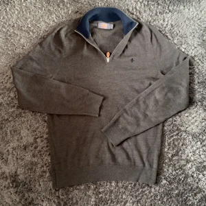 Brun merinoulltröja från Morris - Snygg brun långärmad tröja från Morris i 100% merinoull. Tröjan har en marinblå krage, half zip-dragkedja med läderdetalj och diskret broderad logga på bröstet. Perfekt för lager-på-lager och stilren look.