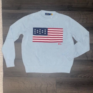 Ralph Lauren stickad tröja  - Ljusblå stickad tröja från Polo Ralph Lauren med amerikansk flagga framtill och RL-broderi. Klassisk rund halsringning och ribbade muddar. Perfekt för dig som vill sticka ut med en ikonisk look. Antingen så är det strl M barn heller så har den blivit tvättat fel för den sitter mer som en XS, så utgå från måtten, längd. 59cm, axelbredd 39cm, längd längst ned på tröjan 40cm. Skicket är helt perfekt knappt använd och inga defekter över huvudtaget!