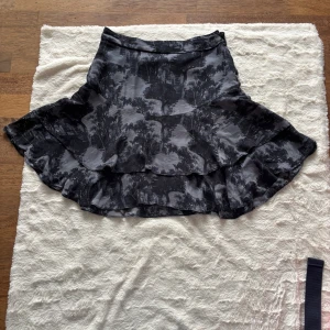 Svart och grå volangkjol från Part Two - Snygg kortkjol från Part Two i svart och grått med abstrakt mönster. Kjolen har volangdetaljer och en lätt glansig yta som ger en cool vibe. Materialet känns mjukt och följsamt, perfekt för dig som gillar att sticka ut med din stil. Svart underkjol. Fint skick 