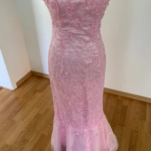 Ljusrosa långklänning med spetsdetaljer - Säljer en elegant ljusrosa långklänning med vackra broderade spetsdetaljer och blommönster. Klänningen har figurnära passform och är tillverkad i ett glansigt material som ger en lyxig känsla. Perfekt för bal eller speciella tillfällen.