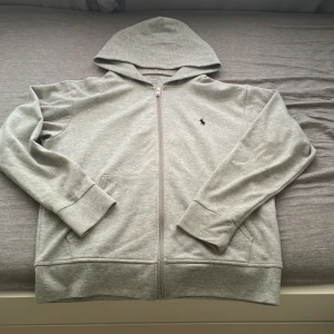 Grå hoodie från Polo Ralph Lauren - Snygg ljusgrå hoodie från Polo Ralph Lauren med dragkedja och klassisk logga på bröstet. Tröjan har huva, två fickor fram och är tillverkad i mjuk bomullsmix. Perfekt för chill dagar eller när du vill ha en clean och enkel stil. Hör av dig vid frågor, mvh Alex 