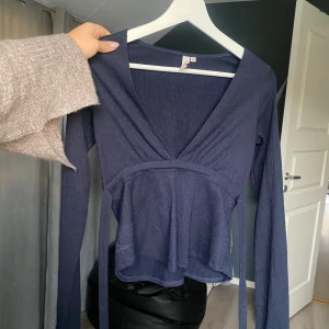 Mörkblå omlottopp med knyt - Supersnygg mörkblå omlottopp med v-ringning och långa ärmar. Toppen har ett knytband i midjan och är tillverkad i ett mjukt, lätt ribbat material. Använd en gång.