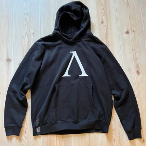 Svart hoodie från Slade Couture XL - Svart hoodie från Slade Couture i storlek XL med stor vit Lambda-symbol på bröstet och tryck med siffran 99 i camouflage på ryggen. Klassisk känguruficka framtill och huva. Perfekt för dig som gillar streetwear och vill sticka ut.
