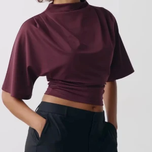 Vinröd croppad polotopp - Snygg vinröd croppad polotopp med korta, vida ärmar och hög hals. Toppens material har en lätt glans och sitter tight runt midjan för en cool siluett. Perfekt för dig som gillar stilrena och trendiga plagg med lite edge.