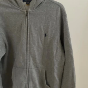 Grå hoodie från Ralph Lauren - Klassisk grå hoodie från Ralph Lauren med dragkedja framtill och broderad logga på bröstet. Tröjan har huva, långa ärmar och två fickor framtill. Perfekt för en chill och stilren look. Storlek Xl barn passar även s 