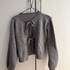 Grå kofta med knyt från SHEIN - Grå stickad kofta från SHEIN i storlek S. Koftan har två snygga knyt framtill och ribbade muddar vid ärmslut och nederkant. Perfekt för lager-på-lager och chill vibes. Materialmix av akryl, polyester och polyamid.
