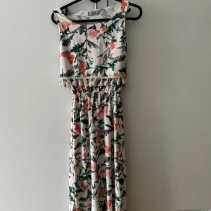 Blommig långklänning från Happy Wippa - Långklänning i vitt med blommigt mönster i grönt och rött från Happy Wippa. Klänningen har ärmlös design, elastisk midja och dekorativa tofsar under bysten. Perfekt för sommarens alla tillfällen med sin luftiga och sköna känsla. Klänningen är liten i storlek. 