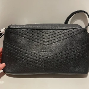 Svart axelväska från Bikkembergs - Snygg svart axelväska från Bikkembergs i skinn med justerbar rem. Väskan har ett stilrent mönster av dekorativa sömmar i V-form och en diskret logga framtill. Perfekt storlek för det viktigaste och levereras med dustbag.