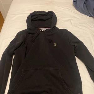 Svart hoodie från U.S. Polo  storlek M - Svart hoodie från U.S. Polo . med broderad logga på bröstet. Tröjan har huva med snörning, känguruficka och ribbade muddar. Skön och stilren modell i mjukt bomullsmaterial, perfekt för chill eller skola.