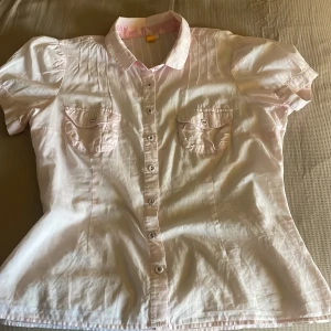 Ljusrosa kortärmad skjorta med fickor - Söt ljusrosa skjorta med korta puffärmar, två bröstfickor med lock och knappar framtill. Skjortan har figursydd passform och fina sömdetaljer upptill. Perfekt för en fräsch och stilren look. Skjortan har ett litet hål i ryggen