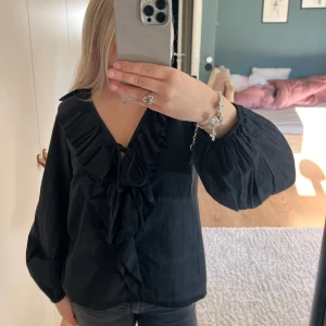 Svart blus med volang från lager 157 - Svart blus från 157 i storlek S med djup v-ringning, stora volanger framtill och knytband vid halsen. Blusen har  ärmar med resår vid handlederna.