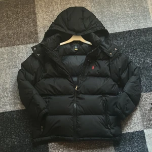 Svart pufferjacka från Polo Ralph Lauren - Säljer en svart pufferjacka från Polo Ralph Lauren med huva och dragkedja. Jackan har två fickor med dragkedja och den klassiska röda Polo-loggan på bröstet. Perfekt för kalla dagar och riktigt snygg till streetwear-stilen. Anledningen att jag säljer den är för att den är ett felköp, jag köpte den och sedan ångrade jag mig, därför säljer jag denna nu, Den är aldrig använd och den är såklar Äkta. Pris kan diskuteras vid snabbt köp