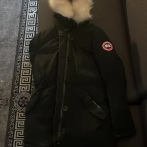 Svart dunjacka från Canada Goose - Säljer en svart dunjacka från Canada Goose med klassisk patch på ärmen och fluffig päls runt huvan. Jackan har dragkedja och knappar framtill, långa ärmar och två snedställda fickor. Perfekt för kalla vinterdagar och riktigt snygg streetstil.