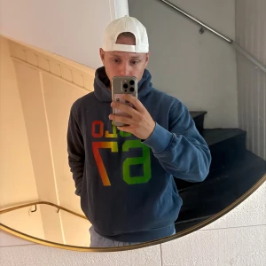Blå hoodie från Polo Ralph Lauren - Säljer en blå hoodie från Polo Ralph Lauren med stor färgglad 67-print i rött, orange, gult och grönt på bröstet. Tröjan har huva, känguruficka och är fodrad med mjuk fleece på insidan. Perfekt för dig som gillar streetwear och vill sticka ut.