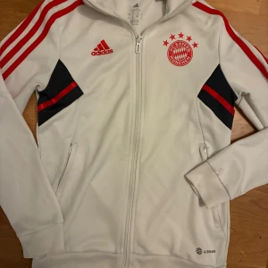 Vit Adidas FC Bayern München tröja - Säljer en vit Adidas tröja med FC Bayern München-logga och röda detaljer. Tröjan har hel dragkedja, ståkrage, röda ränder på ärmarna och svarta paneler med röda inslag. Perfekt för fotbollsfans som vill sticka ut.