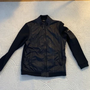 Svart bomberjacka från Plein Sport - Svart bomberjacka från Plein Sport med snyggt tryck på ryggen och vargdetalj. Jackan har dragkedja framtill, ribbade muddar och blandade material – glansig polyester på framsidan och mjukare tyg på ärmar och rygg. Perfekt för dig som gillar sportig och urban stil.