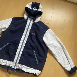  vindjacka Peak Performance - Snygg tvåfärgad vindjacka från Peak Performance i marinblått och vitt. Jackan har huva med broderad logga, dragkedja framtill, två fickor med blå dragkedjor och justerbar nederkant. 