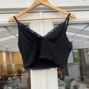 Svart croppad linnetopp med spets - Snygg svart croppad linnetopp med tunna axelband och spetsdetaljer längs både urringning och nederkant. Toppen har omlott-effekt framtill och är tillverkad i ett mjukt manchestertyg som ger en cool känsla.