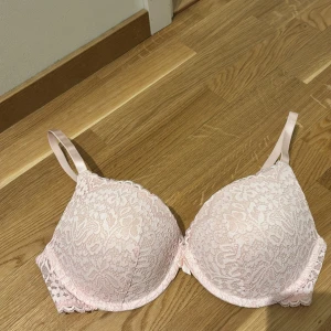Ljusrosa spets-bh med blommönster - Söt ljusrosa bh med vadderade kupor och vackert blommigt spetsmönster. Axelbanden är justerbara och det finns en liten rosett mellan kuporna. Perfekt för dig som gillar romantiska detaljer och mjuka pastellfärger.