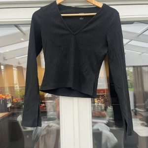 Svart ribbad långärmad topp - Svart långärmad topp från med ribbad struktur och v-ringning. Modellen är croppad och har en smickrande passform som sitter snyggt på kroppen. Perfekt att styla med jeans eller kjol för en trendig look.