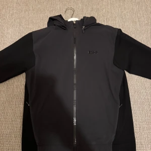  BOSS vindjacka - Svart vindjacka från BOSS med huva och hel dragkedja framtill där BOSS-loggan syns längs dragkedjan och på bröstet.                            Storlek L