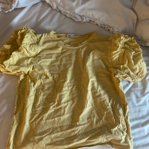 Gul topp med puffärmar från Zara - Superfin gul topp från Zara med korta puffärmar som ger en trendig och lekfull look. T-shirten är i mjuk bomull och har rund halsringning. Perfekt för dig som vill sticka ut med färg och volym på ärmarna.