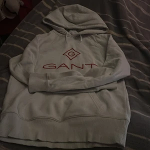 Vit hoodie från GANT med röd logga - Vit hoodie från GANT med stor röd broderad logga på bröstet. Klassisk modell med huva och snörning, samt ribbade muddar vid ärmslut och nederkant. Mjuk och skön, perfekt för chill dagar eller när du vill ha en clean look.