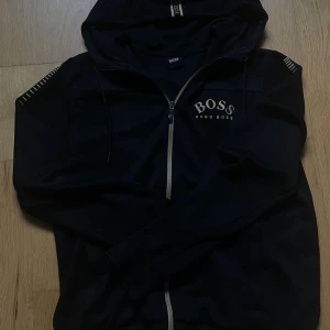 Svart hoodie från Hugo Boss med dragkedja - Svart hoodie från Hugo Boss med vit logga på bröstet och reflecterande dragkedja. Tröjan har hel dragkedja, huva med snörning och är tillverkad i mjuk bomull. Perfekt för dig som gillar stilrena och sportiga plagg.