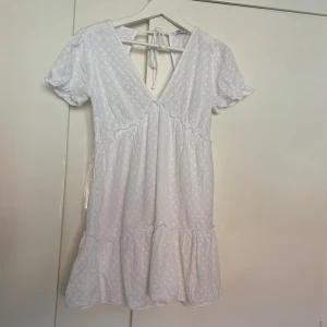 Vit kort klänning från Zara med knyt - Superfin vit kort klänning från Zara i bomull med broderade detaljer och v-ringning. Puffiga korta ärmar, volang nertill och knyt i ryggen.