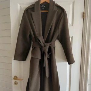 Brun kappa från Zara - Stilren khaki grön kappa från Zara med bälte i midjan och stora fickor framtill. Endast använd en gång och säljer pågrund av att det var fel storlek! Jag skulle säga att det är en väldigt mörk grön färg och kan förvirras med brun men i solljus är den grön!