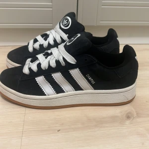 Adidas Campus svarta sneakers - Svarta Adidas Campus sneakers med klassiska vita ränder på sidorna, vit logga på plös och häl samt breda vita skosnören. Ovandel i mocka och sula i gummi med brun undersida. Snygg retrostil som funkar till det mesta.