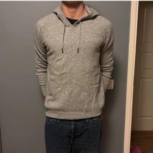 Grå stickad hoodie - Mysig grå stickad hoodie med huva och snörning. Tröjan har lång ärm och en klassisk passform som funkar till allt. Den är i bra skick då den är använd ett fåtal gånger bara.