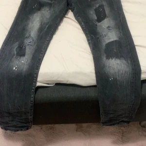Svarta Dsquared2 jeans med slitningar - Säljer ett par svarta jeans från Dsquared2 med coola slitningar och färgstänk på benen. Jeansen har en tvättad look och klassisk femficksdesign. Märkeslogga på bakfickan och vid gylfen. Perfekta för dig som gillar streetstyle och unika detaljer.