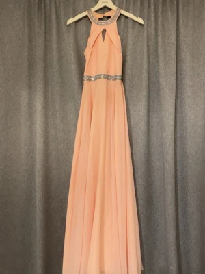 Ljusrosa långklänning med strass - Elegant ljusrosa långklänning med glittrande strassdetaljer vid halsringning och midja. Klänningen har ärmlös design, hög hals och ett snyggt nyckelhålsutskärning fram. Flödande kjol i lätt material som ger en drömmig look. Perfekt att ha på bal eller bröllop. Storleken är barn storlek, 14 år. Men passar för äldre personer också. Den är använd endast en gång.