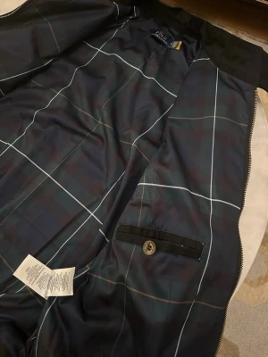 Svart bomberjacka från Polo Ralph Lauren -                                                  Våga skriv pris förslag   Svart bomberjacka från Polo Ralph Lauren med röd broderad logga på bröstet. Jackan har dragkedja framtill, två fickor med knappstängning och ståkrage med dubbla knappar. Insidan har ett snyggt rutigt foder i mörkblå toner och en innerficka med knapp.