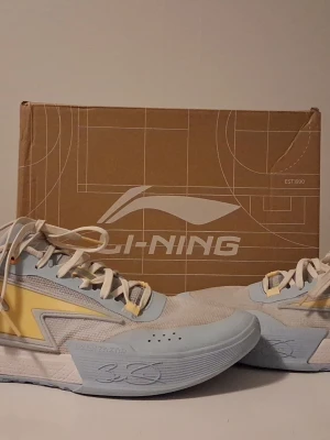 Li-Ning sneakers i pastellblå och gul - Säljer ett par Li-Ning sneakers i fräsch pastellblå och gul färg med vita detaljer. Skorna har snörning, mesh och syntetmaterial, samt coolt blixtmönster på sidan. Broderad text 'Hustle' på hälen och signaturdetaljer på sulan. Perfekta för dig som vill sticka ut.du får även med dig sko lådan.