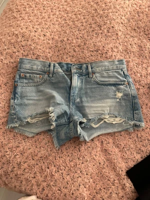Ljusblå denimshorts med slitningar - Säljer ett par ljusblå denimshorts från Perfect Jeans med råa, fransiga kanter och slitningar framtill. Klassisk femficksmodell med bälteshällor och knappstängning. Perfekta för chill dagar och sommarhäng.