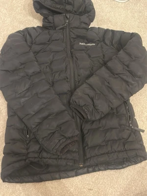 Svart pufferjacka Peak Performance - Svart pufferjacka från Peak Performance med huva och dragkedja framtill. Jackan har quiltad design och två sidofickor med dragkedja. Perfekt för kalla dagar och har en stilren look som funkar till det mesta. 2 små hål som är lagade på jackan, därav priset . Storlek 160 (xs)